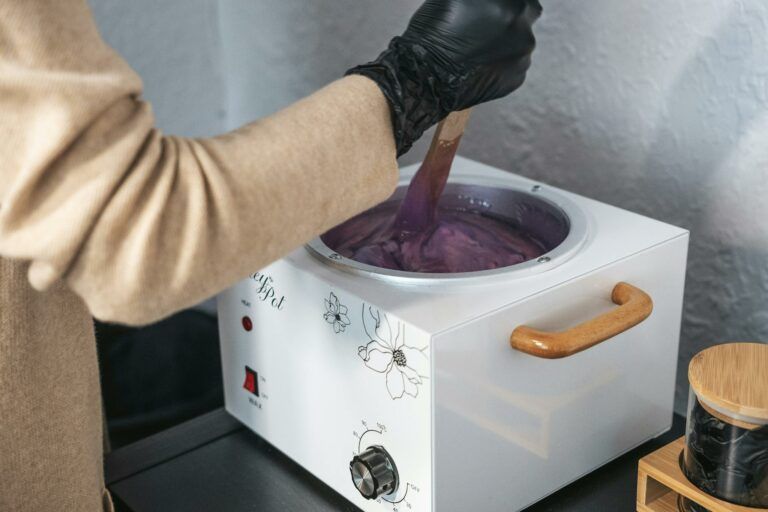 An esthetician wearing gloves stirring purple wax in a wax pot indoors.